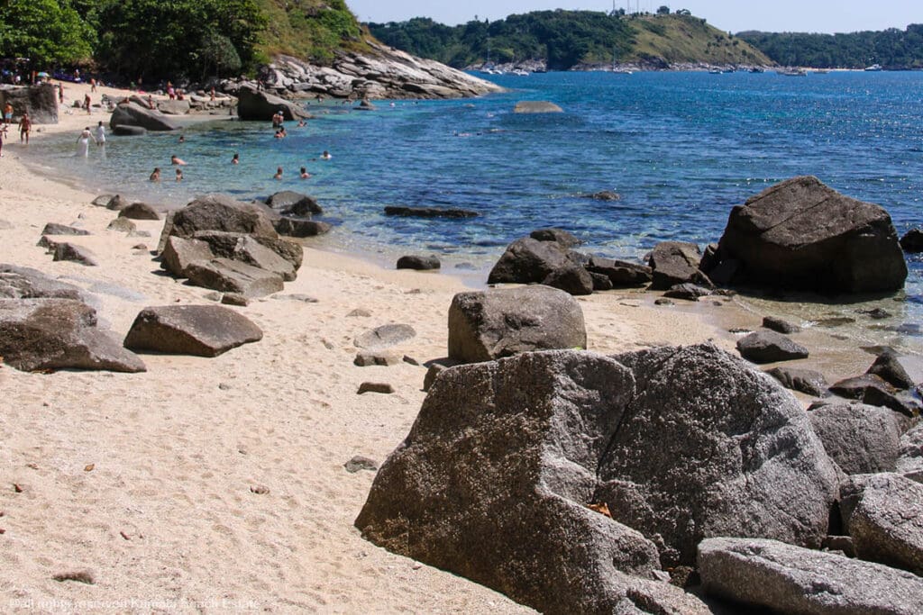 Beachgoers at Ao Sane Beach in Phuket, with rocky shores, clear blue waters, and lush greenery in the background.