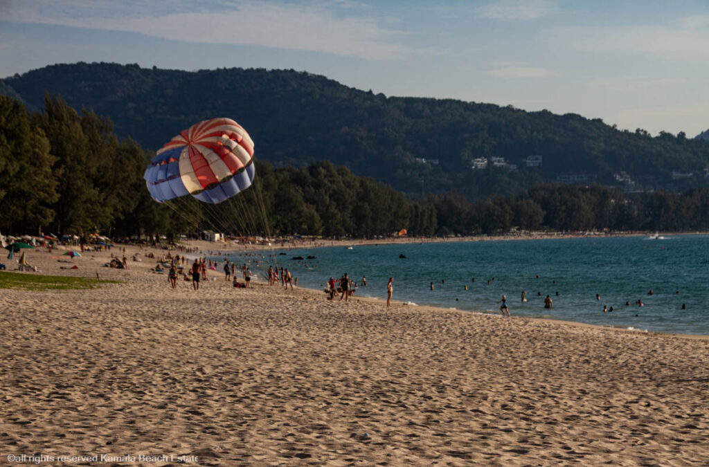 Parasailing at Bang Tao Beach, Phuket with beachgoers enjoying the sun and sea, and lush green hills in the background.