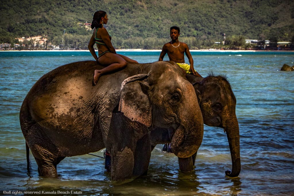 Two people riding elephants in the water at Hua Beach with lush greenery in the background.