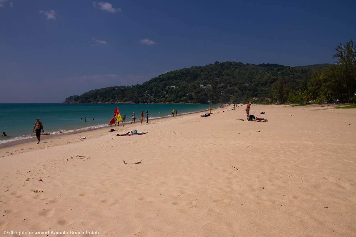 A vibrant view of Karon Beach in Phuket, featuring golden sand, turquoise waters, and a lively atmosphere with sunbathers and beachgoers enjoying the serene surroundings.