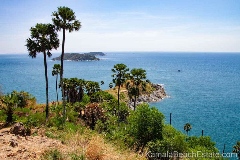 View of Promthep Cape in Phuket, Thailand, featuring a rocky headland, palm trees, and the turquoise waters of the Andaman Sea under a clear sky.