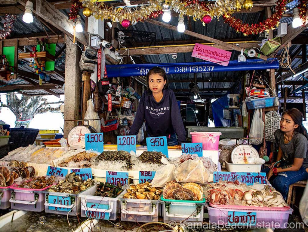 Seafood stall at Rawai Seafood Market in Phuket, Thailand, displaying a variety of fresh seafood including scallops, lobster, and fish with price tags. Two local vendors stand behind the counter offering their daily catch.